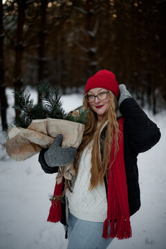 Smiling young woman in winter outfit holding pine branches outdoors in snowy forest. Concept of Christmas holidays, winter lifestyle, happiness, seasonal nature and festive mood.