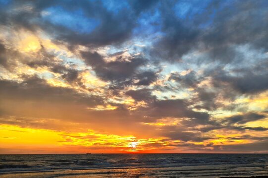 Dramatic ocean sunset featuring fiery orange and yellow sky contrast with dark blue clouds over gentle sea waves.