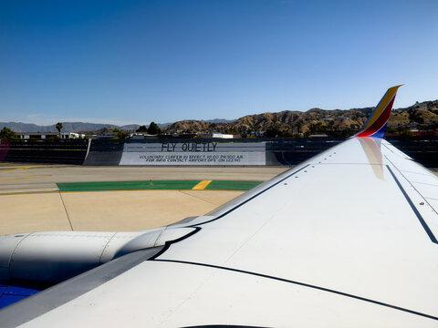 BURBANK, CA, APR 28, 2026: Southwest Airlines wingtip and engine beside airport runway and taxiway, with a "Fly Quietly" curfew sign and hills in the distance