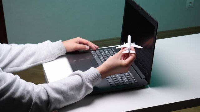 Person's hands typing on a laptop keyboard while holding a small white airplane model, symbolizing planning for flight reservations and holiday trips using technology