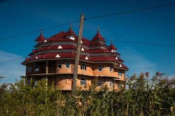 Unfinished eccentric mansion in rural Romania with multiple red turret roofs and exposed brick walls, surrounded by tall wild grass under a clear blue sky. Kitschy architecture. © Anze