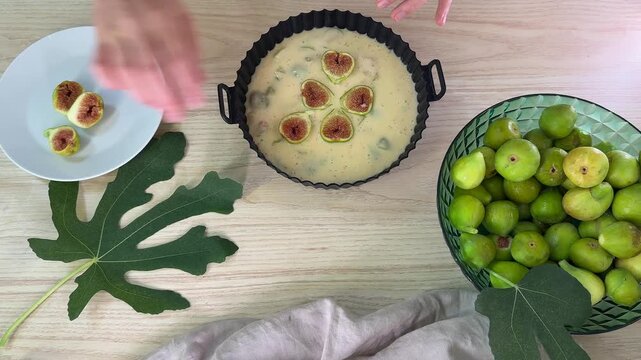 A person places sliced figs into a cake batter inside a black fluted baking pan, surrounded by a bowl of green figs and a fig leaf on a light wooden surface