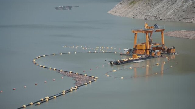 Reservoir dredging operation, floating dredger removes sediment near rocky dam slope, supporting water infrastructure, maintenance, engineering, environmental cleanup.