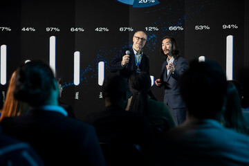 Obraz premium Two presenters of different ages deliver a speech at a business conference while professionals attend an international summit in an auditorium.