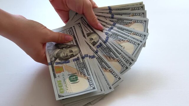 A bundle of 100 dollar notes is reviewed one by one before a planned expense. The scene suggests checking available funds, verifying the amount and preparing cash for a significant purchase.