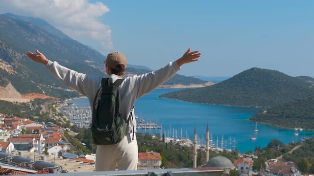 Tourist enjoys the view of a coastal town. Young female traveler with a backpack standing on a viewpoint, raising her arms in joy while admiring the beautiful panoramic view of the coastal town