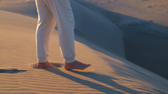Woman's bare feet walking on a desert sand dune at sunset. Young woman's bare feet walking in slow motion along the crest of a sand dune, leaving footprints in the warm, textured sand during a sunset