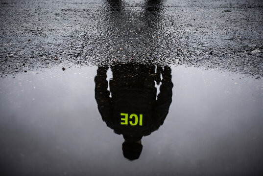 Conceptual reflection of an ICE officer in a water puddle on wet asphalt street pavement symbolizing immigration enforcement law and government surveillance
