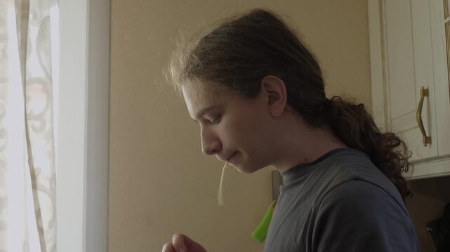 Medium close profile of teenage boy eats cheese, standing beside window in warm muted home interior, mouth slightly open, hair tied back in ponytail