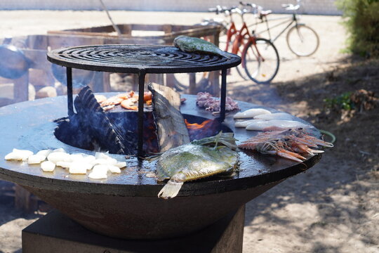 Fresh seafood including fish, langoustines, shrimp, and cuttlefish cooking on a large round outdoor brazier plancha grill over open wood fire on &Icirc;le d'Aix,