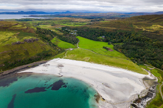 Calgary Bay on Isle of Mull, Scotland, white shell sand beach bordering turquoise water below green pastures, woodland, and rolling coastal hills, aerial view