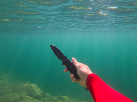 First-person underwater photograph showing a diver's hand in a red sleeve holding a black fixed-blade diving knife in clear green water.