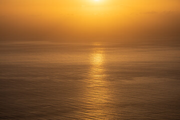 Obraz premium Golden sunset over calm ocean water with bright sun reflection and haze background
