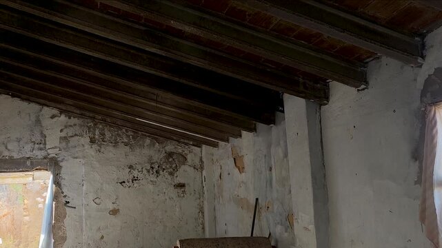 Interior view of an old abandoned ruined building with decayed wooden ceiling beams, peeling plaster walls, and a improvised window curtain