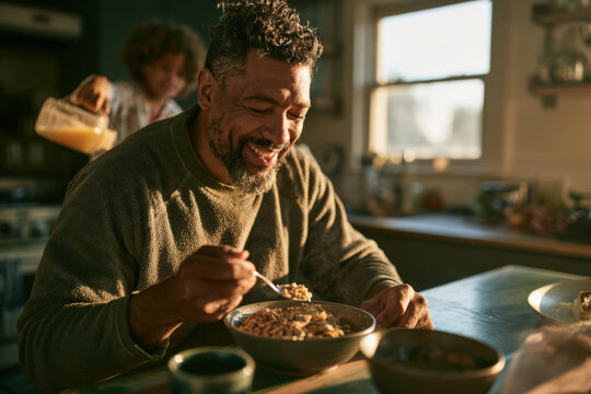 Man home from a night shift eats breakfast at a small kitchen table at first light while a child in pajamas pours juice, plain uncluttered kitchen, soft copy space. Tired but happy to be home.