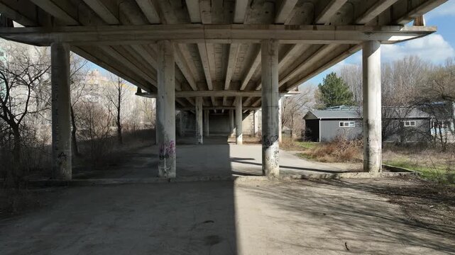 Forward movement beneath a concrete highway overpass reveals pillars, shadows, and a sloped embankment. Urban infrastructure, underpass corridor, transportation decay