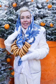 Smiling woman in traditional Russian shawl holding string bag with mandarins and sushki by snowy Christmas tree
