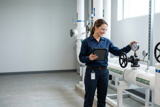 A smiling woman technician in safety glasses inspects a chilled water valve in a clean data center cooling plant room. Skilled trades and technology workforce concept.