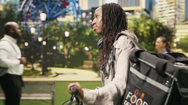 Woman bicycle courier stands with her bike and insulated delivery backpack in a city park, ready to continue food delivery work after checking the next route as an immigrant gig worker.