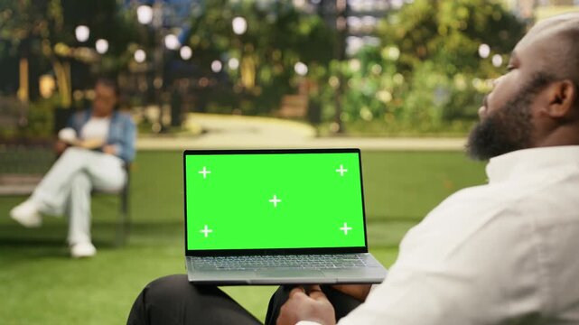 Businessman holds a laptop with a green screen chroma key in an urban park, providing an editable widescreen display for advertising or business content.