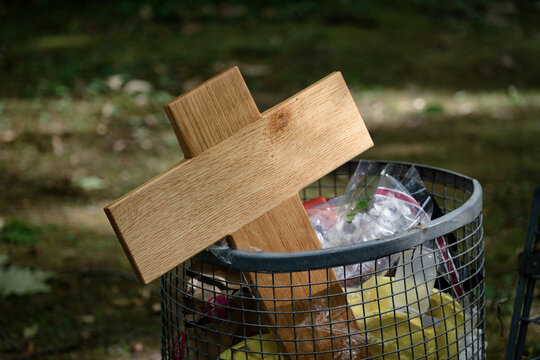 A simple wooden cross disposed of in an outdoor wire mesh garbage can along with plastic waste