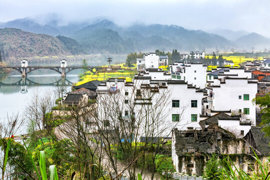 A traditional village with white-walled houses by a river, backed by misty mountains and yellow rapeseed fields.