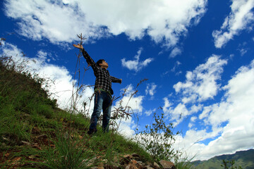 An adult stands on a grassy hillside with arms outstretched, looking up at a bright blue sky with white clouds, surrounded by natural terrain and vegetation. © tai