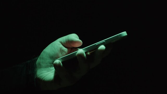Phone at night, Close-up macro hands of a man scroll news feed on mobile phone app application. Screen of the mobile phone illuminates male hand. 