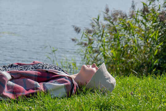 Woman in a light sun hat and checked shirt lying on green grass beside the water with her face turned toward sunlight. Outdoor relaxation and peaceful nature escape