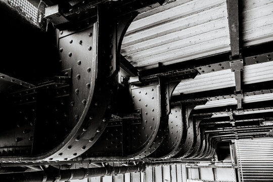 Berlin, Germany - July 5, 2026: Black and white close-up beneath a historic riveted steel bridge reveals repeating curved girders, metal plates and structural connections.