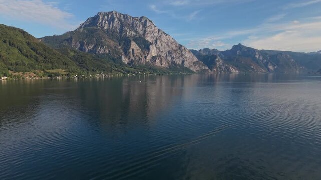 Beautiful aerial drone flight over the calm blue waters of Lake Traunsee in Austria, revealing dramatic rocky Alpine mountains, forested slopes and small lakeside settlements on a clear summer day.