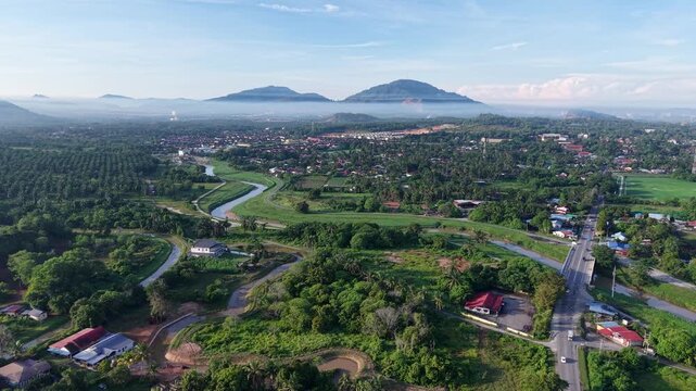 Green valley meanders through village, framed by distant hills. Mist curls around peaks at Perai, Penang