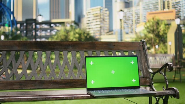 Isolated green screen laptop sits on a wooden bench in a city park, framed as an establishing urban landscape shot with a front-facing chroma key display for screen replacement use.