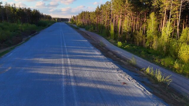 An overhead overpass with an offset overpass over a dirt road surface being prepared for asphalting, surrounded by a pine forest, on a sunny summer evening.