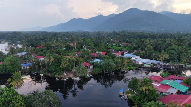 Drone panorama of tropical lakeside village with palm trees, boats and misty mountain backdrop at dusk at Tasik Raban