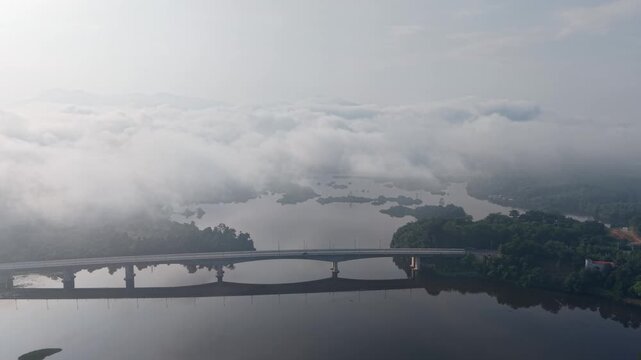 A serene aerial view of a calm river with a long bridge crossing it, surrounded by misty fog and lush greenery at Tasik Raban