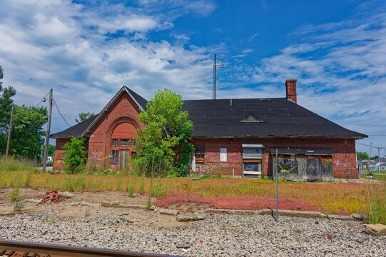 An abandoned Pennsylvania Railroad red brick train station with boarded windows, an overgrown platform and adjacent railroad tracks beneath a bright blue sky. Plymouth, Indiana Evoking decay, history 