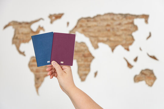 Hand holding two biometric passport covers, blue and burgundy, against a blurred carved world map on a wall