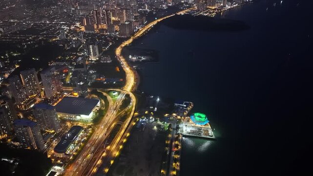 Drone aerial night shot of coastal city highway curving past illuminated waterfront gallery and urban skyline at Gelugor, Penang