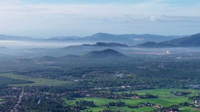 Aerial view shows misty mountain ranges. Distant valleys display vibrant green fields at Kubang Semang