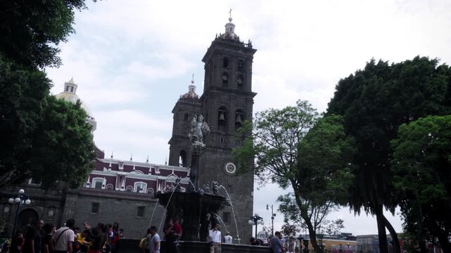 Archangel San Miguel fountain in central zocalo with Puebla Cathedral towers in background framed by green trees on cloudy day