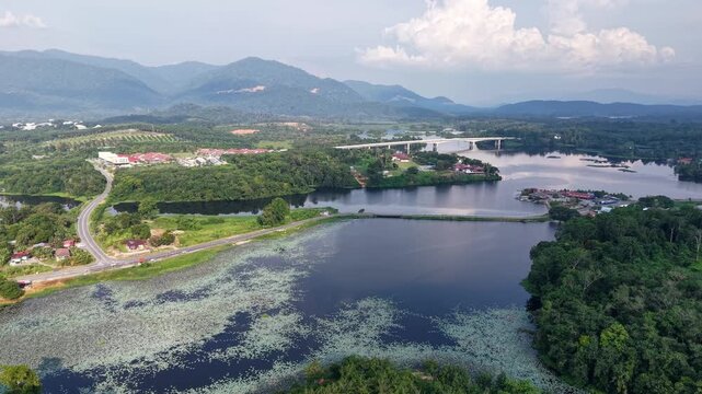 A breathtaking aerial view of a wide river surrounded by dense green forest with a bridge and winding road nearby under a partly cloudy sky and distant mountains at Tasik Raban