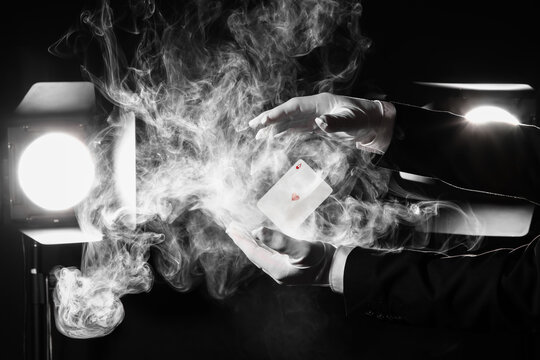 Male magicians hands with levitating playing card and smoke against glowing lights on dark background