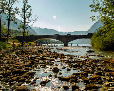 People enjoy a sunny day swimming in the clear waters near a stone bridge over Lake Bohinj in Slovenia, surrounded by scenic mountains and a rocky riverbank, creating a peaceful outdoor leisure