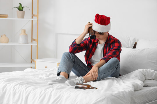 Young man with hangover and Santa hat putting bottle to his head in bedroom after Christmas party
