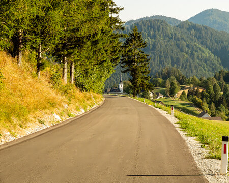 A winding asphalt road leads through a lush, forested mountain landscape towards a small church and village in the distance, capturing the serene beauty of the rural Alpine countryside.