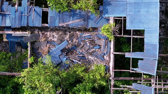Aerial view shows collapsed blue corrugated roofs. Scattered metal sheets cover the ground