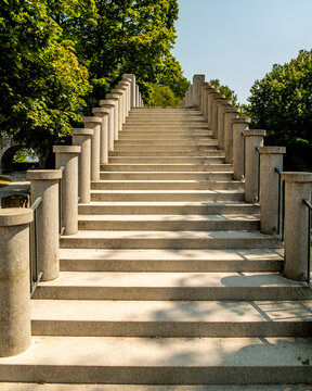 A symmetrical stone staircase flanked by columns leads upwards towards lush greenery in a park, serving as an architectural path for pedestrians to reach a higher level or viewpoint.