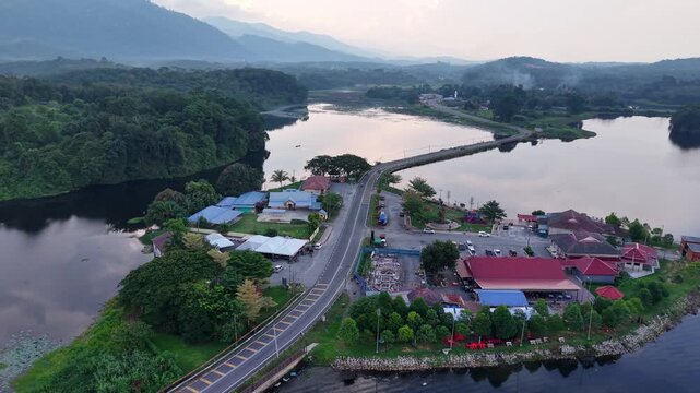 Aerial view of a countryside road passing over a bridge surrounded by a calm river and lush green forest with scattered houses under a cloudy sky at Tasik Raban