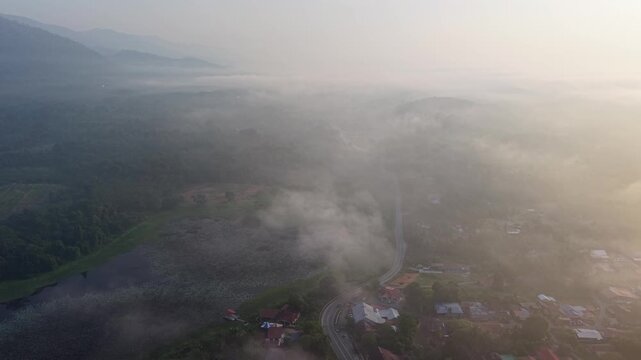 Golden hour light softens mountain peaks. Fog clings to green fields and winding road at Tasik Raban